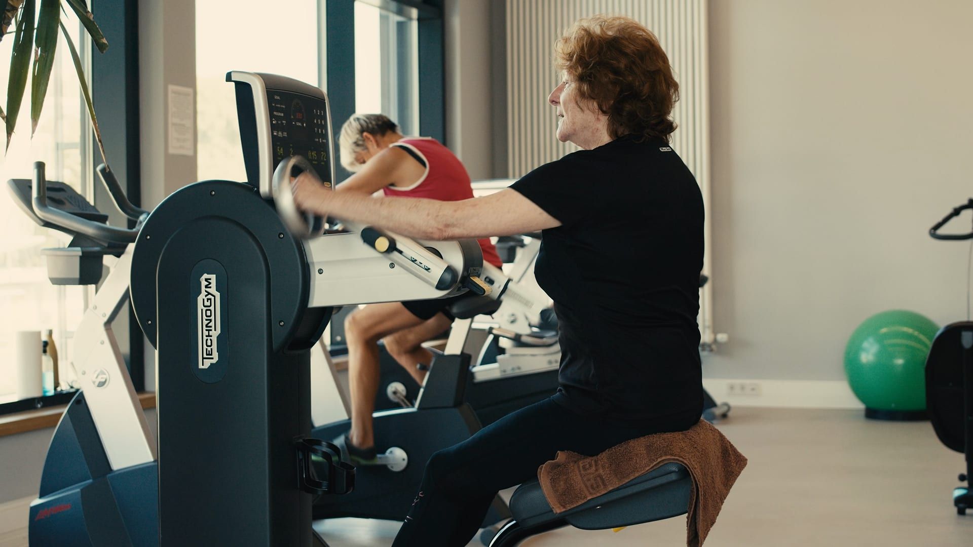 Eine Frau trainiert an einem Ergometer auf der Trainingsfläche, im Hintergrund eine weitere Person an einem Gerät am Fenster.